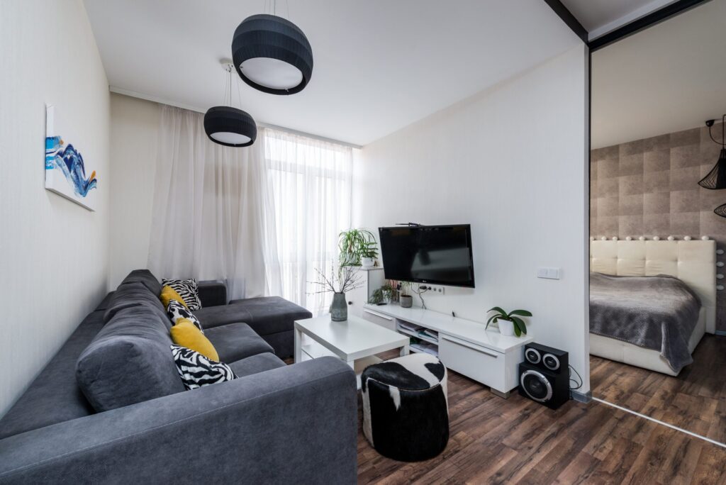 Nowoczesny salon z szaro-czarną sofą, telewizorem i roślinami, z widokiem na sypialnię przez przeszklone drzwi